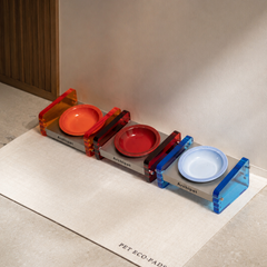 Triple connected colorful acrylic cat bowl stand, orange red blue elevated neck-protect pet bowl with ceramic bowls on beige PET ECO-PADS mat, modern aesthetic pet product photo