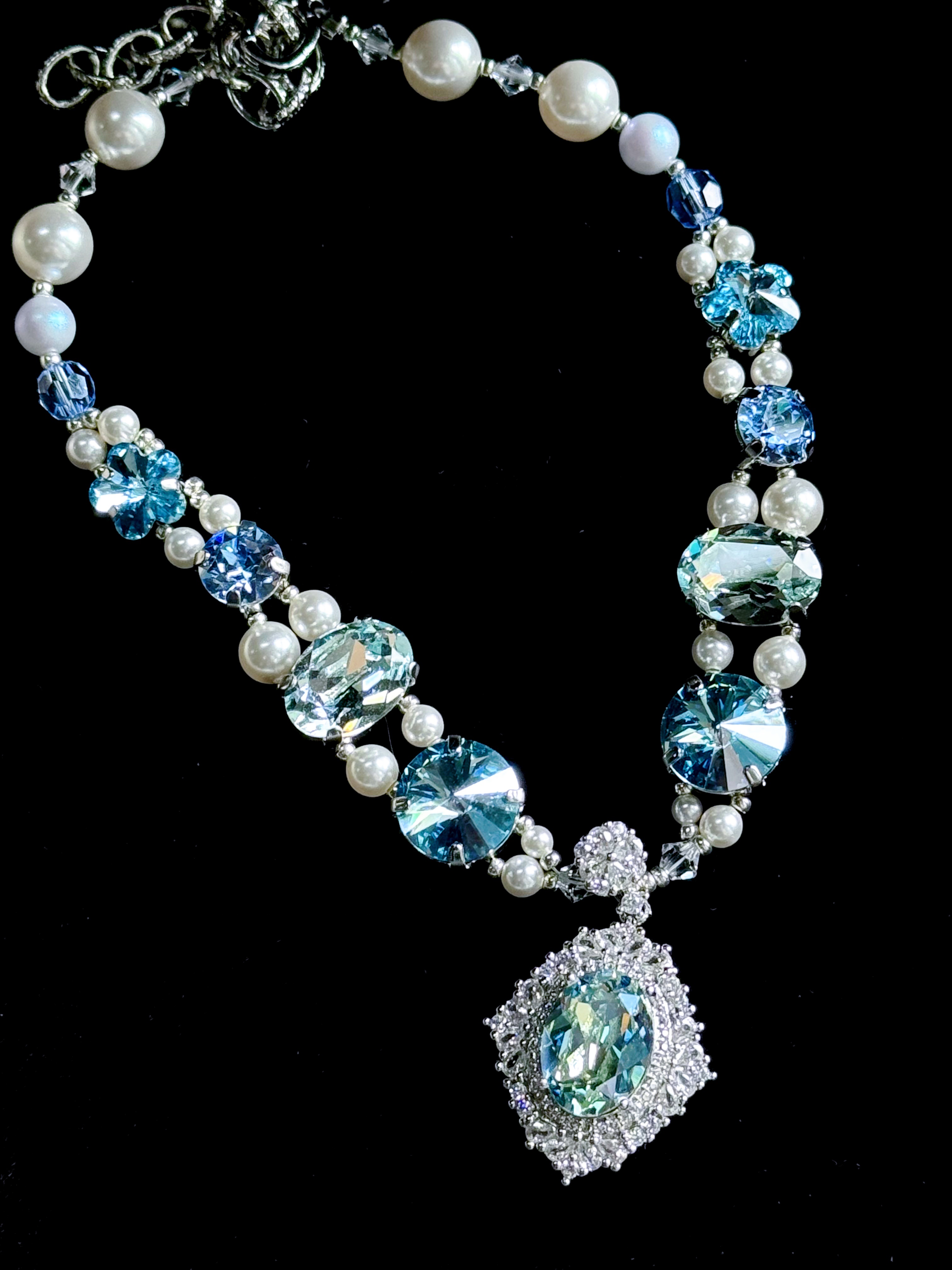 Full shot of elegant dog necklace with layered blue crystals, flower shaped gems, white pearls and big aquamarine pendant, black background flat lay