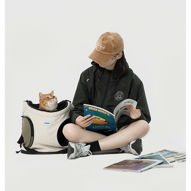 floor-placed-pet-front-backpack-carrier-cat, Pet front backpack carrier placed on ground, cat inside bag, woman reading magazine beside portable pet travel bag